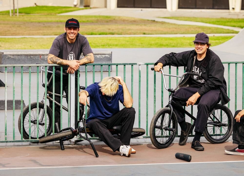 Trzech mężczyzn siedzi na rowerach BMX przy metalowym ogrodzeniu na skateparku. Na ziemi leży fioletowy głośnik mobilny.