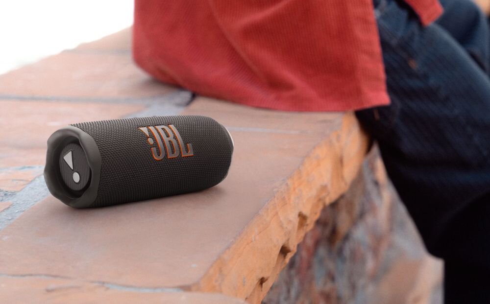Na obrazie widoczny jest przenośny głośnik z logo \'JBL\', leżący na kamiennej powierzchni. W tle znajduje się osoba ubrana w czerwony materiał.