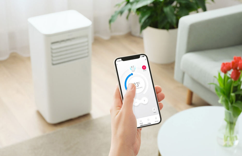 Na obrazie widać białe urządzenie klimatyzacyjne oraz dłoń trzymającą smartfon z aplikacją do sterowania klimatyzatorem. W tle znajduje się sofa, rośliny i stolik z wazonem czerwonych kwiatów.