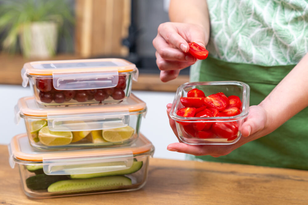 Na obrazie widoczne są cztery przezroczyste pojemniki na żywność z pomarańczowymi pokrywkami, wypełnione różnymi produktami spożywczymi, takimi jak pomidory, winogrona, cytryny i ogórki. Osoba trzyma jeden z pojemników z pokrojonymi pomidorami.
