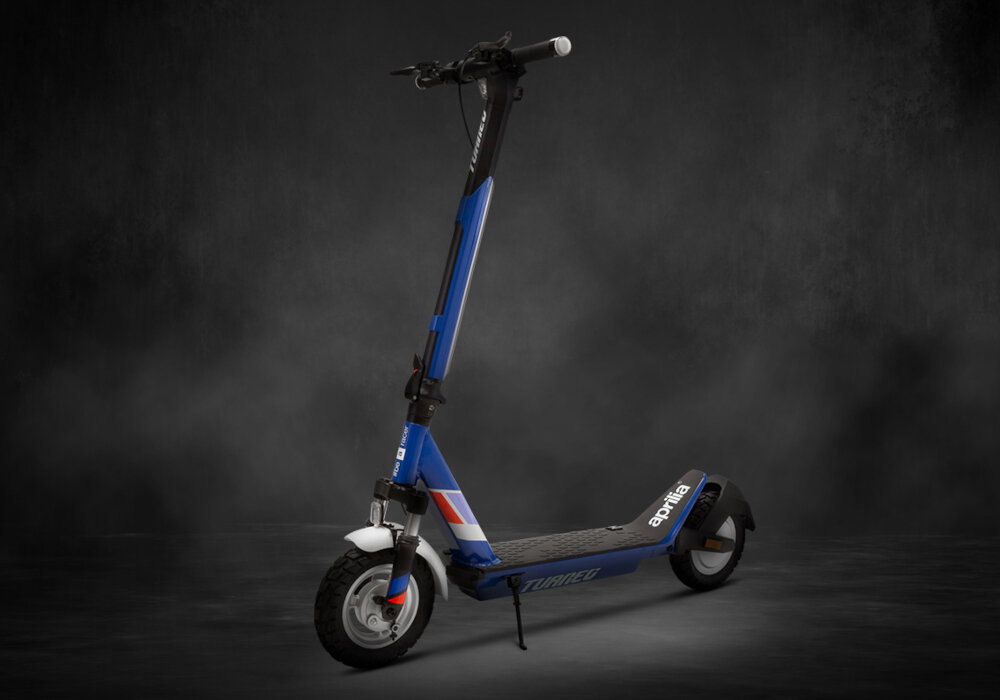 Granatowa hulajnoga elektryczna z białymi i czerwonymi akcentami, widoczna na ciemnym tle. Na podeście napis \'Tuareg\', a na tylnym błotniku logo \'aprilia\'.