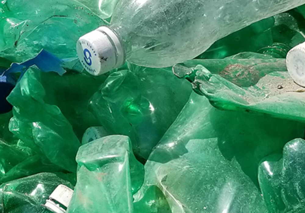 Na obrazie widoczne są zgniecione zielone i przezroczyste plastikowe butelki. Na jednej z nakrętek znajduje się niebieski symbol z literą \'S\'.