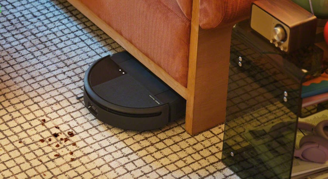 Czarny robot sprzątający znajduje się pod kanapą na wzorzystym dywanie. Obok robota widoczne są ciemne plamy na dywanie.