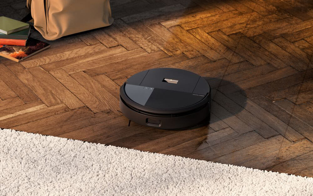 Czarny robot sprzątający znajduje się na drewnianej podłodze obok jasnego dywanu. W tle widoczna jest torba i kilka książek.