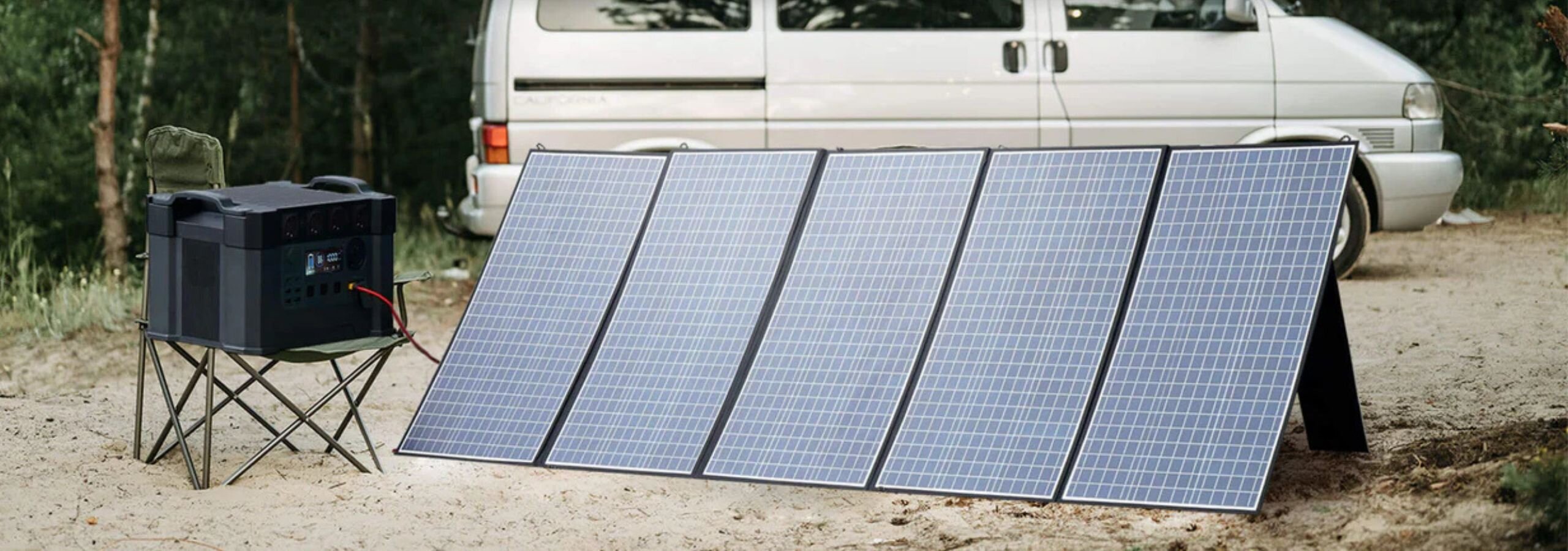 Na obrazie widoczny jest składany panel solarny złożony z pięciu segmentów, ustawiony na ziemi obok białego vana. Obok panelu stoi przenośne urządzenie na składanym stoliku.