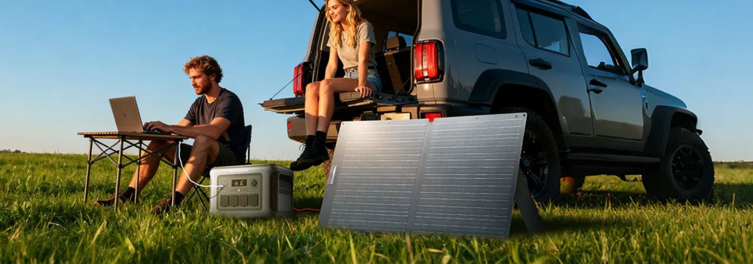 Na trawie stoi składany panel solarny obok samochodu terenowego, przy którym siedzą dwie osoby. Obok panelu znajduje się przenośne urządzenie z wyświetlaczem.