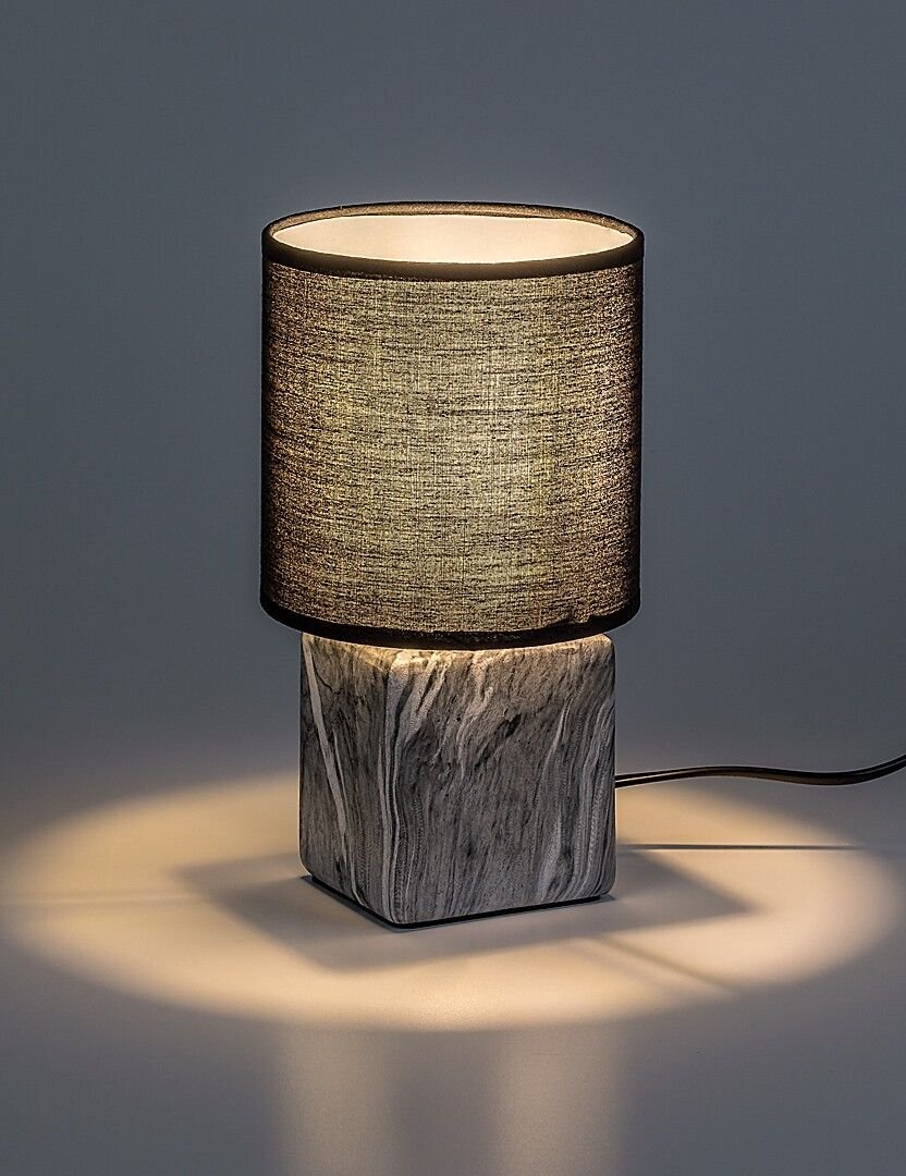 Lampa nocna z cylindrycznym abażurem w ciemnym kolorze i podstawą imitującą marmur. Światło tworzy delikatny efekt na powierzchni, na której stoi lampa.