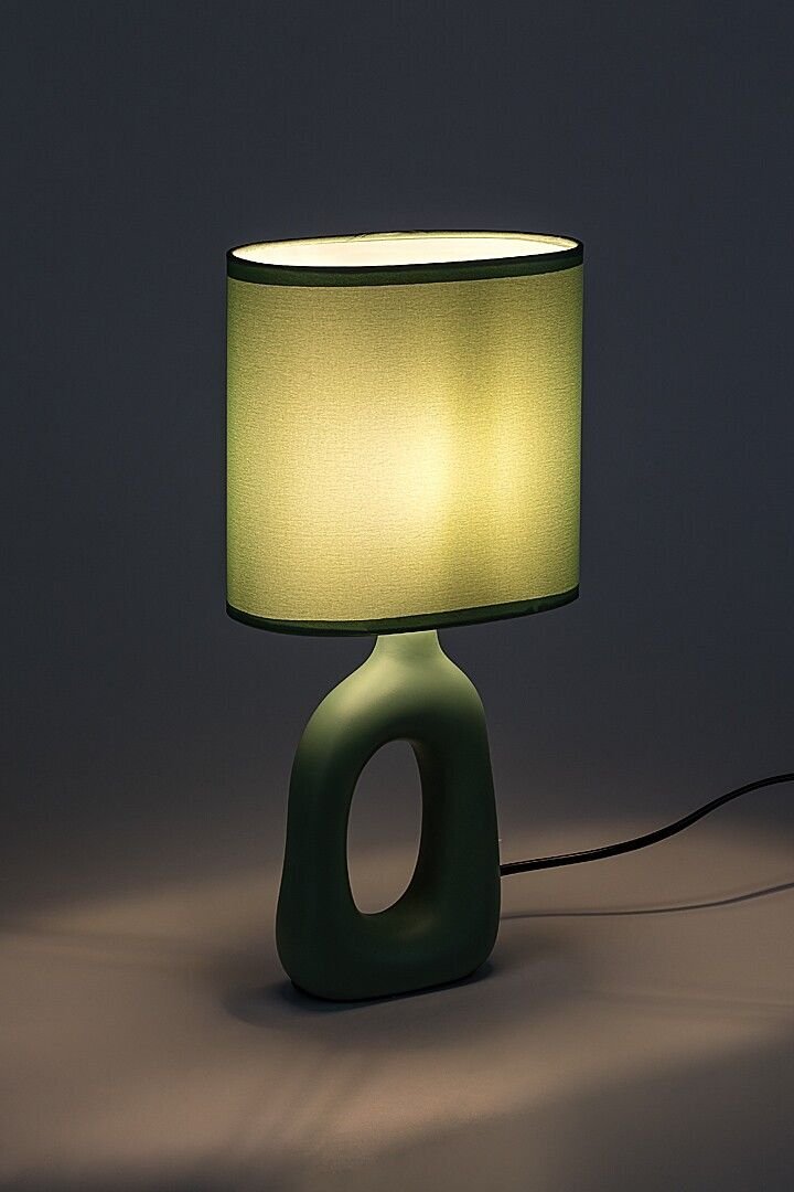 Stylowa lampa nocna z zielonym, owalnym kloszem i podstawą o nowoczesnym kształcie z otworem. Lampa emituje ciepłe, przytulne światło.