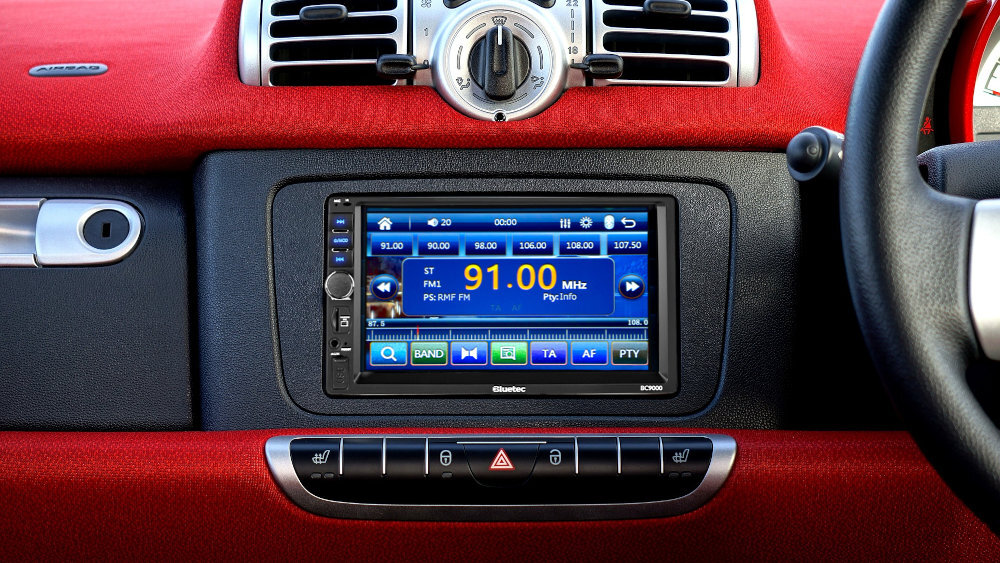 Radio samochodowe z wyświetlaczem pokazującym częstotliwość 91.00 MHz oraz różne opcje sterowania. Na dole ekranu widoczny jest napis \'Bluetec BC 9000\'.