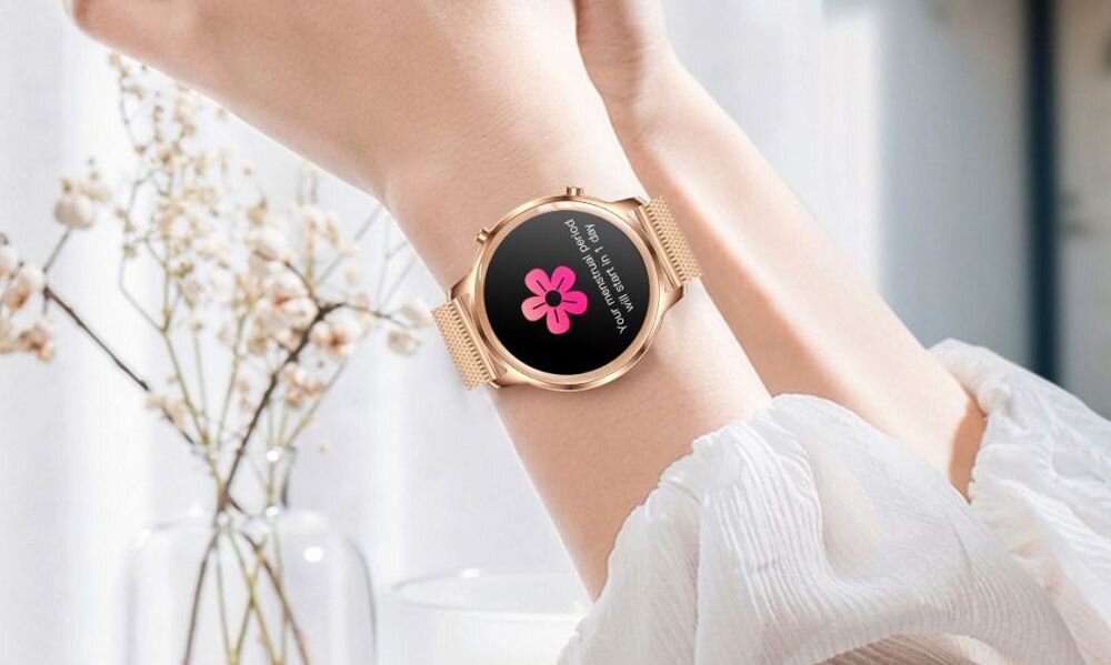 Osoba nosi smartwatch z okrągłym, złotym kopertą i czarnym wyświetlaczem, na którym widoczny jest różowy kwiat oraz tekst. W tle znajdują się rozmyte gałązki z białymi kwiatami w wazonie.