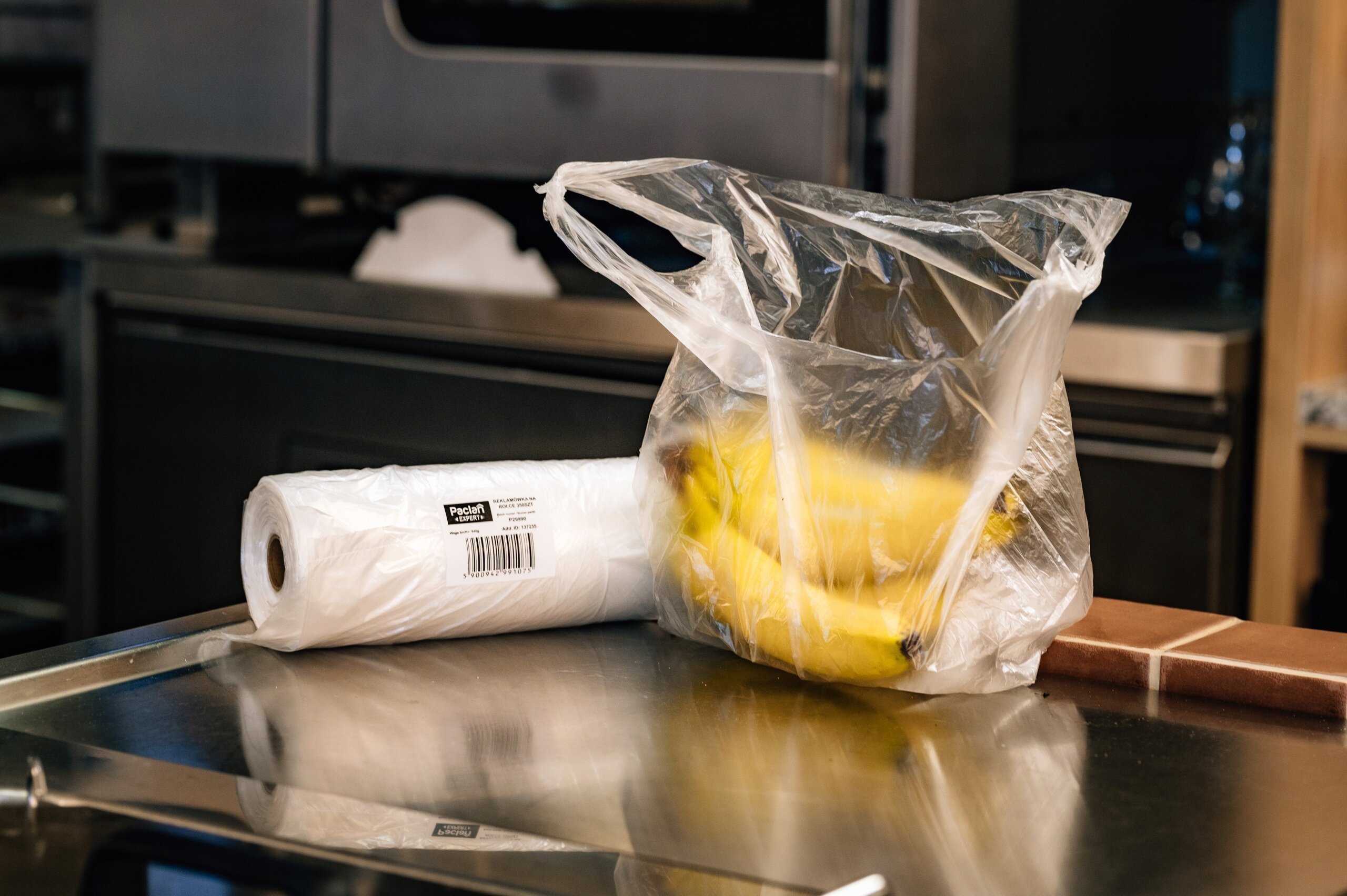 Na blacie kuchennym znajduje się przezroczysta reklamówka z bananami oraz rolka białych worków z widocznym logo \'Paclan\'.