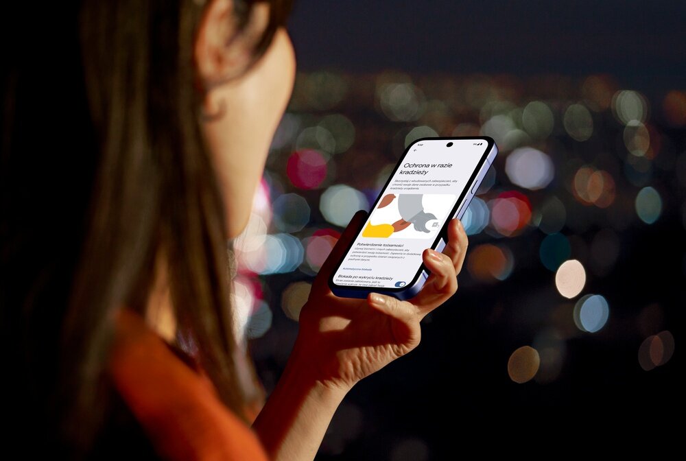 Osoba trzyma smartfon, na ekranie widoczny jest tekst i kolorowa grafika. W tle rozmyte światła miasta nocą.