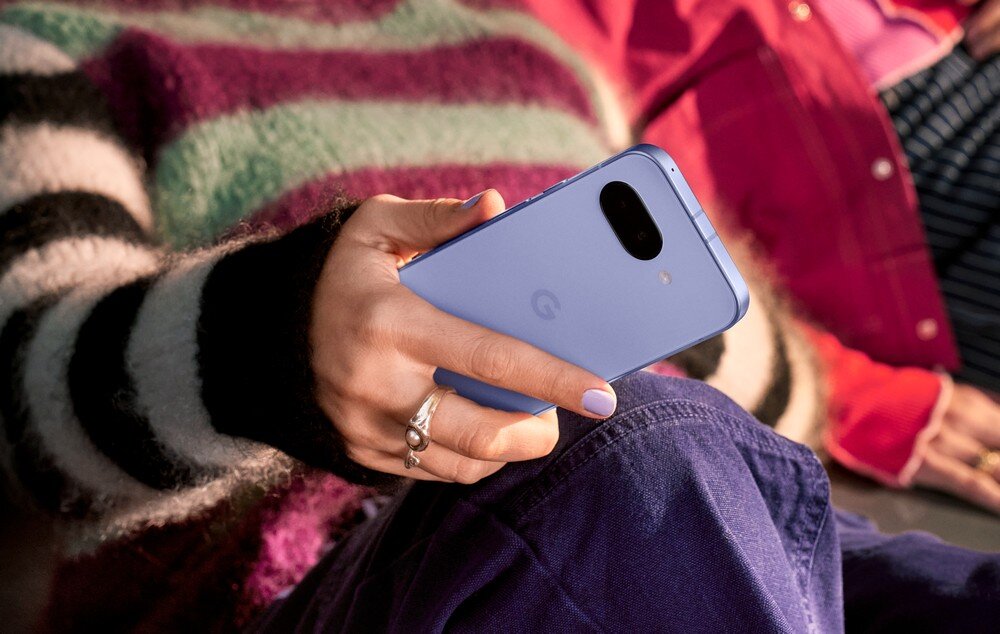 Osoba trzyma niebieski smartfon z logo \'G\' na tylnej obudowie. W tle widoczne są kolorowe ubrania.