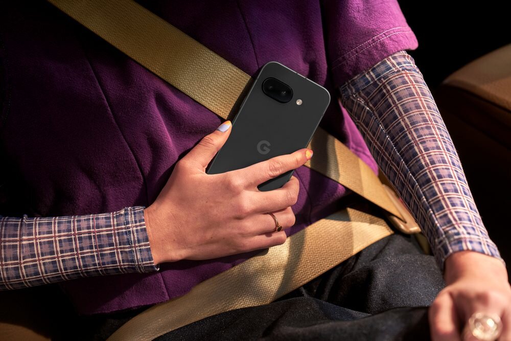 Osoba trzyma czarny smartfon z logo \'G\' na tylnej obudowie. Ubrana jest w fioletową koszulkę i ma zapięty beżowy pas bezpieczeństwa.