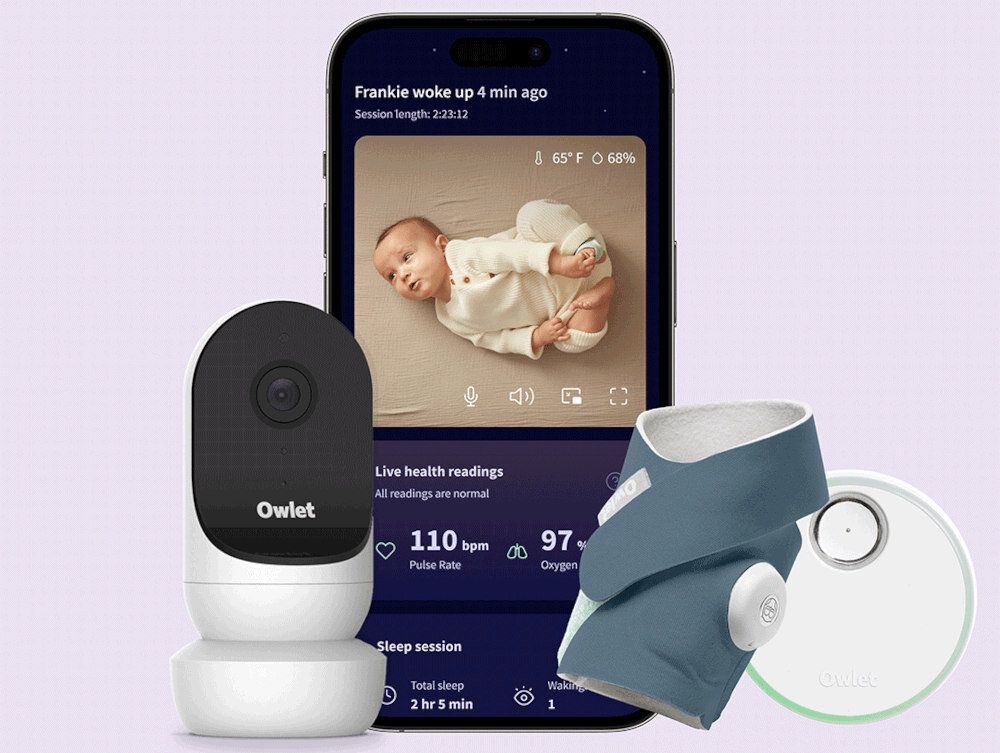 Na obrazie widoczna jest elektroniczna niania z kamerą Owlet oraz monitor oddechu w kolorze granatowym. W tle znajduje się smartfon z aplikacją pokazującą obraz dziecka i dane zdrowotne, takie jak puls i poziom tlenu.