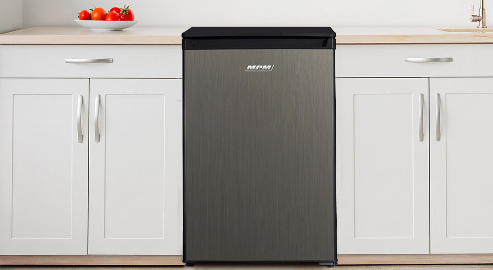 Lodówka w kolorze inox z czarnym blatem, z widocznym logo MPM, stoi pomiędzy białymi szafkami kuchennymi. Na blacie obok znajduje się miska z pomidorami.