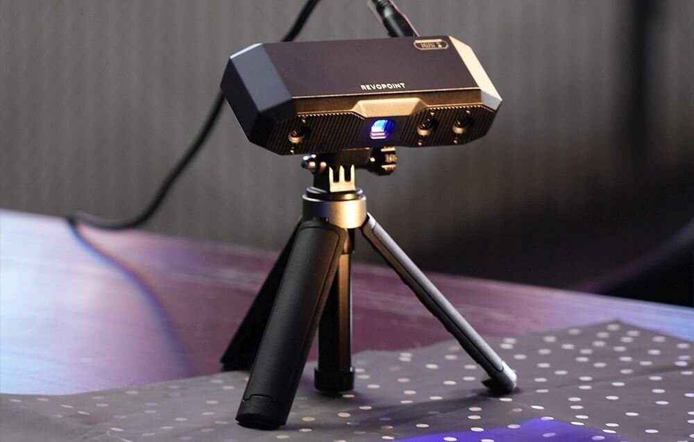 Skaner 3D Revopoint Mini Mini z niebieskim światłem na trójnogu na biurku z matą w kropki