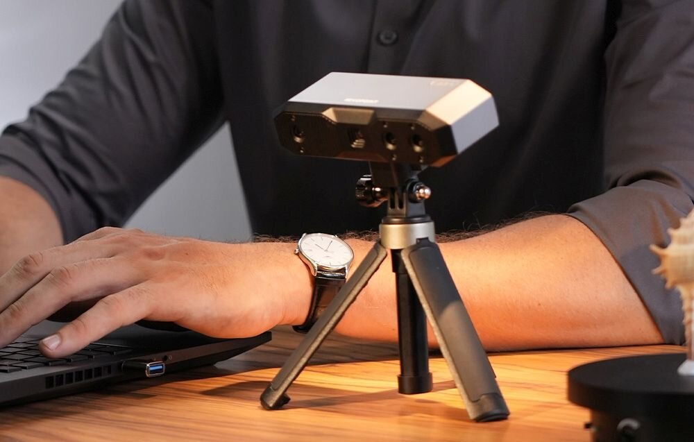Skaner 3D Revopoint Mini Mini z niebieskim światłem na trójnogu na biurku z matą w kropki