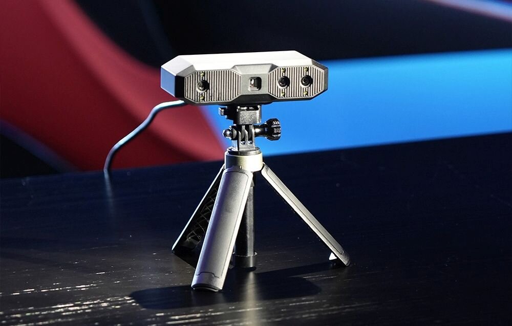 Skaner 3D Revopoint Mini Mini z niebieskim światłem na trójnogu na biurku z matą w kropki
