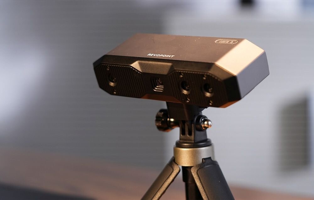 Skaner 3D Revopoint Mini Mini z niebieskim światłem na trójnogu na biurku z matą w kropki