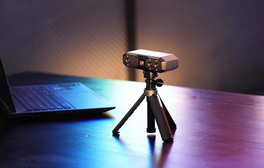Skaner 3D Revopoint Mini Mini z niebieskim światłem na trójnogu na biurku z matą w kropki