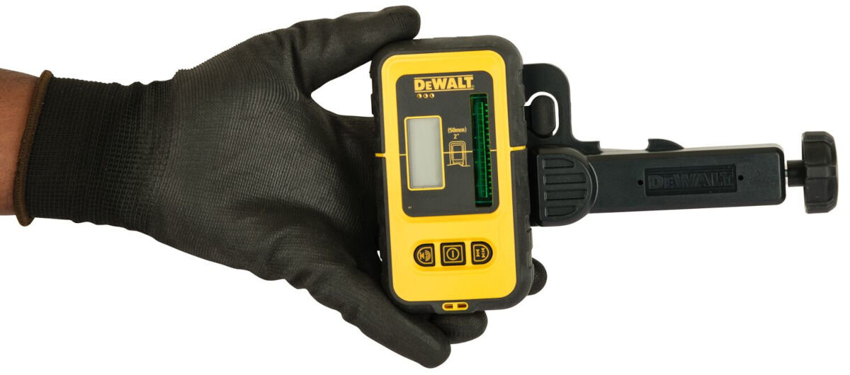 Osoba w czarnej rękawicy trzyma detektor wiązki laserowej z logo DEWALT na żółtej obudowie. Na urządzeniu widoczne są przyciski i wyświetlacz z zielonym wskaźnikiem.