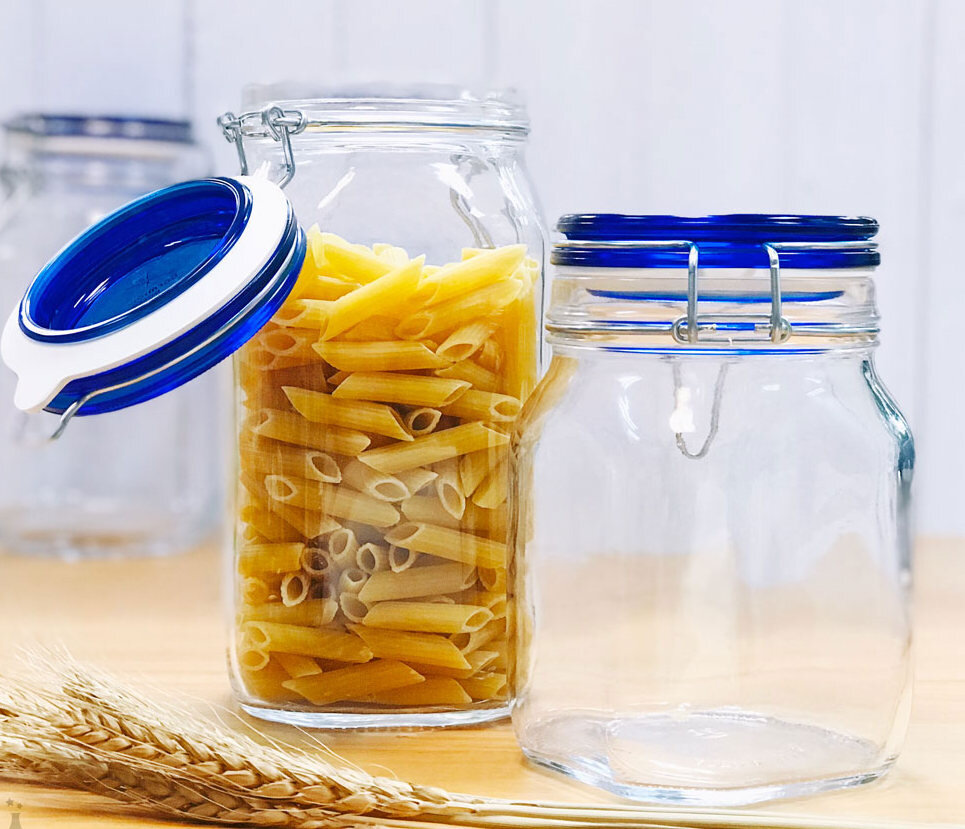 Na obrazie znajdują się dwa szklane słoiki hermetyczne z klipsem, jeden z nich wypełniony jest makaronem penne. Słoiki mają niebieskie pokrywki, a w tle widoczne są kłosy zboża.