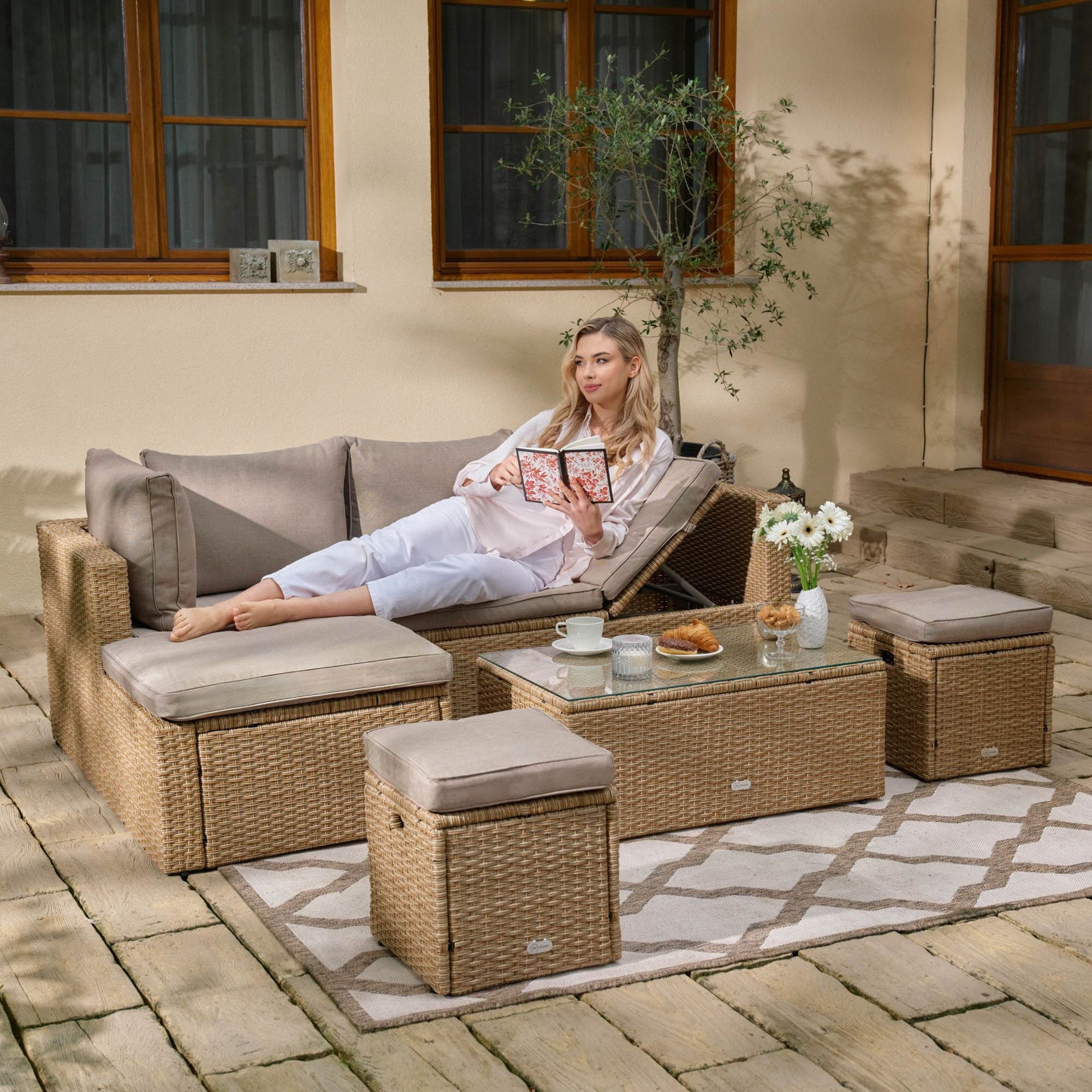 Na tarasie znajduje się zestaw mebli ogrodowych z beżowymi poduszkami, w tym sofa, stolik i pufy. Na stoliku leżą filiżanki, talerzyk z rogalikiem i wazon z kwiatami.