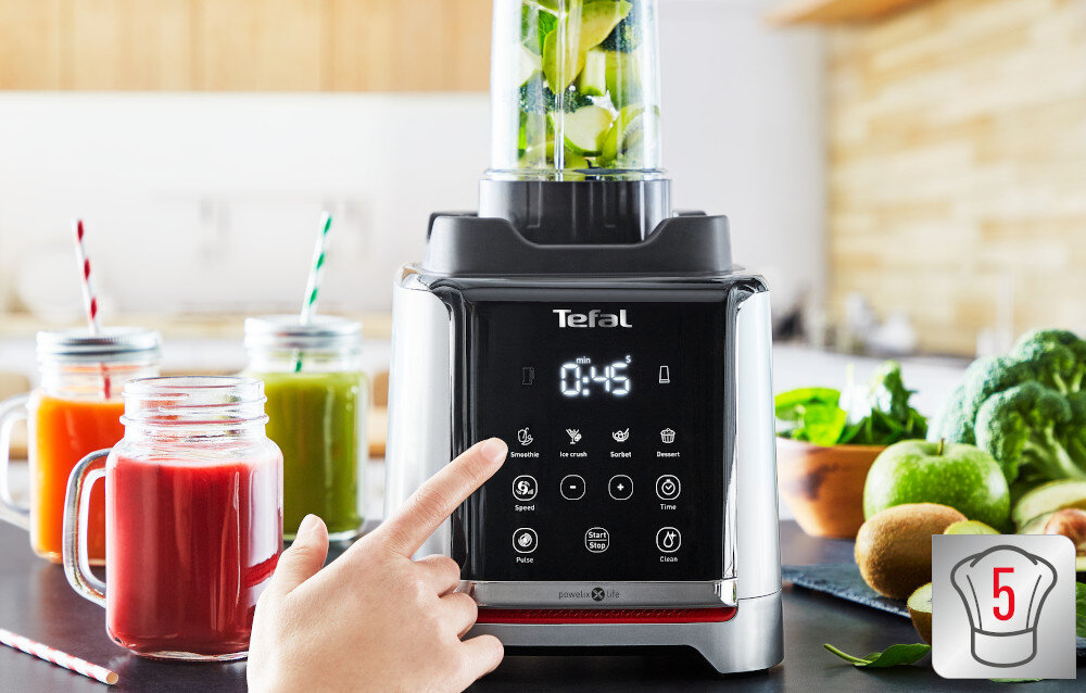 Na obrazie widoczny jest blender kielichowy z logo Tefal, z wyświetlaczem pokazującym czas 0:45 oraz różnymi ikonami funkcji. W tle znajdują się trzy szklanki z kolorowymi napojami i warzywa na blacie.