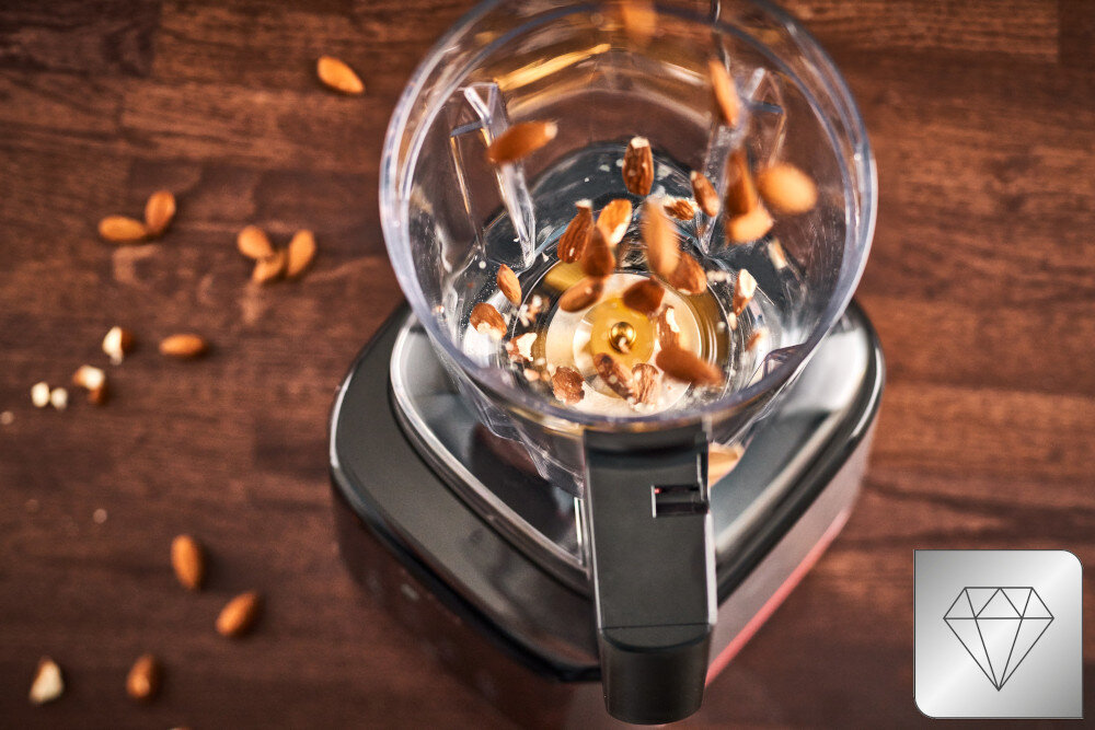 Na obrazie widać blender kielichowy z migdałami w środku, umieszczony na drewnianej powierzchni. W prawym dolnym rogu znajduje się ikona diamentu.