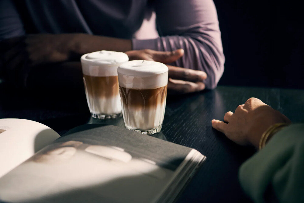 Na stole znajdują się dwa szklane kubki z kawą latte macchiato, obok leży otwarta książka. W tle widoczne są ręce dwóch osób.