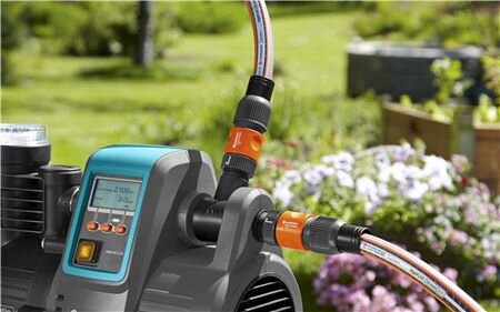 Na obrazie widoczny jest hydrofor z wyświetlaczem LCD, na którym widać cyfry. W tle znajduje się ogród z kwiatami i zielenią.
