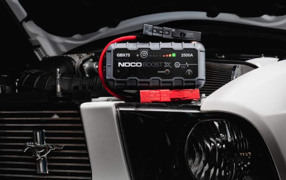 Na obrazie widoczne jest urządzenie rozruchowe NOCO Boost X GBX75 umieszczone na samochodzie, z widocznymi czerwonymi i czarnymi kablami. Na urządzeniu znajdują się napisy: \'GBX75\', \'NOCO BOOST X\', \'2500A\'.