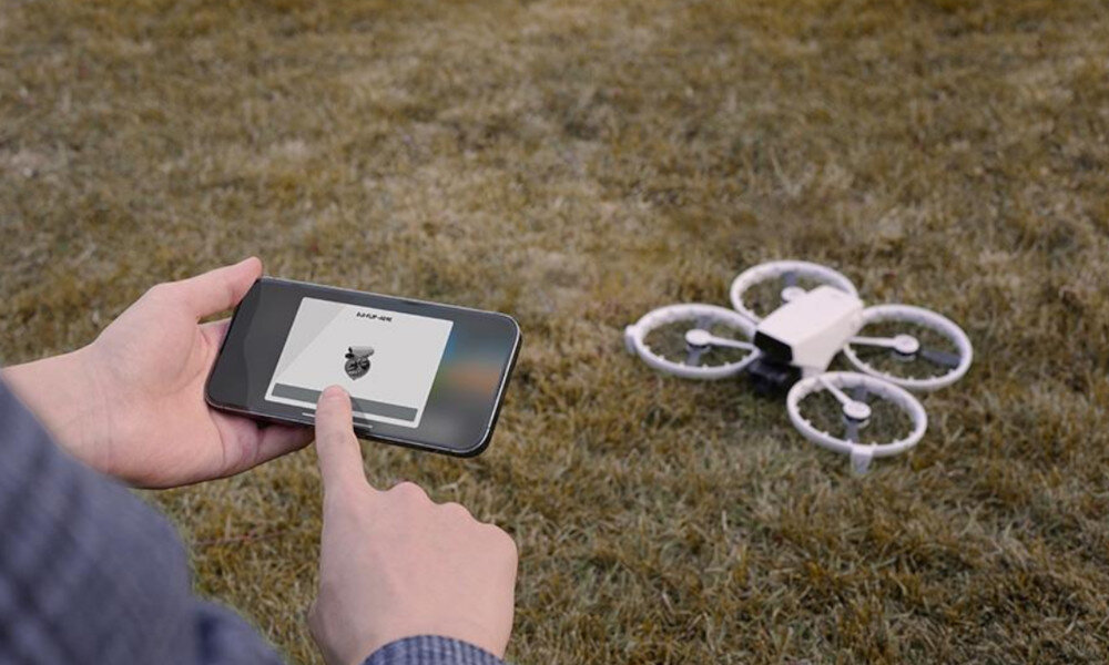 Osoba trzyma smartfon z wyświetlonym obrazem drona i tekstem \'DJI RC2\', a na trawie widać drona z czterema śmigłami.