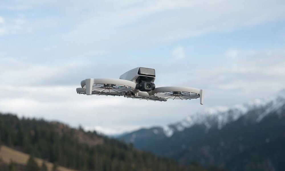 Dron w locie na tle górskiego krajobrazu z widocznymi drzewami i zaśnieżonymi szczytami. Dron ma kompaktową konstrukcję z kamerą z przodu.