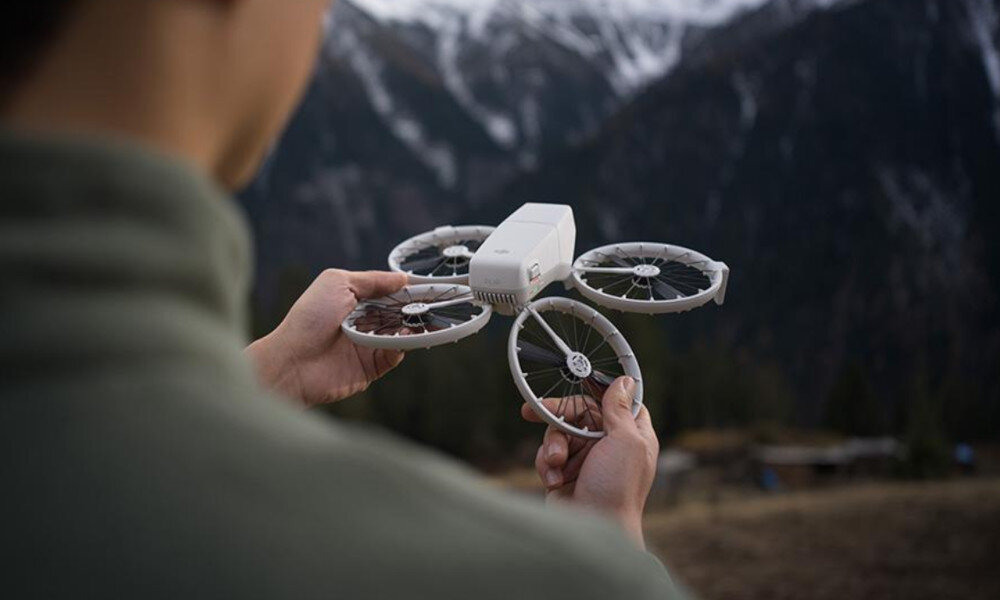 Osoba trzyma drona z czterema wirnikami na tle górskiego krajobrazu. Dron jest biały i ma kompaktową konstrukcję.