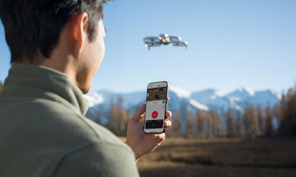 Osoba trzyma smartfon z aplikacją do sterowania dronem, w tle widać latającego drona oraz górski krajobraz.
