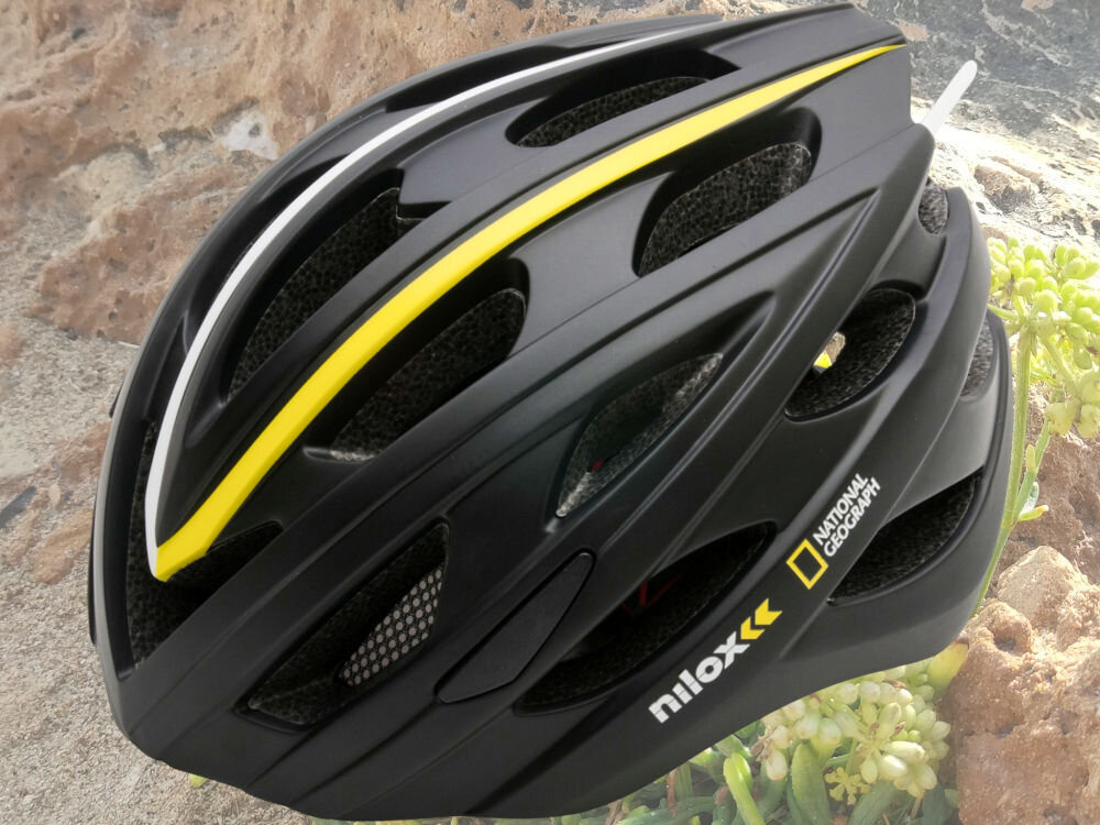 Czarny kask z żółtymi i białymi akcentami, z widocznymi napisami \'nilox\' i \'National Geographic\'. Kask posiada otwory wentylacyjne i znajduje się na tle skał i roślinności.