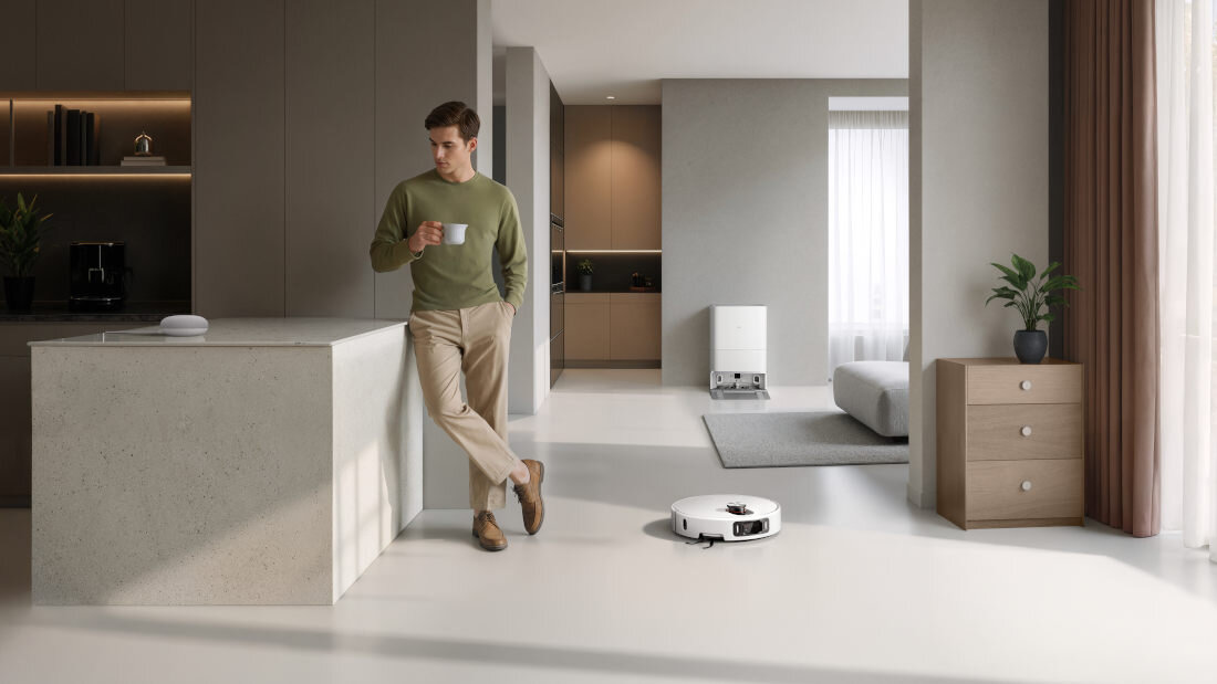 W nowoczesnym wnętrzu kuchni stoi mężczyzna z kubkiem, a na podłodze znajduje się biały robot sprzątający. W tle widoczna jest część salonu z rośliną na komodzie.