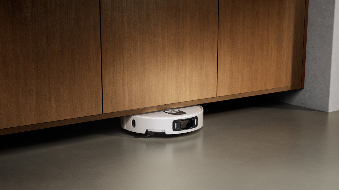 Biały robot sprzątający znajduje się pod drewnianą szafką na gładkiej podłodze.