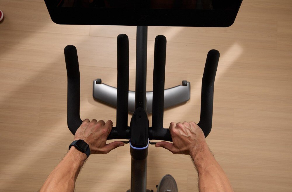 Osoba trzyma kierownicę roweru treningowego z wyświetlaczem na drewnianej podłodze. Na nadgarstku widoczny jest zegarek sportowy.