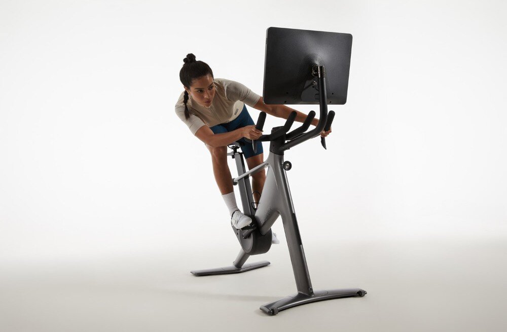 Osoba korzysta z roweru treningowego z dużym ekranem zamontowanym z przodu. Rower ma nowoczesny design i jest ustawiony na jasnym tle.