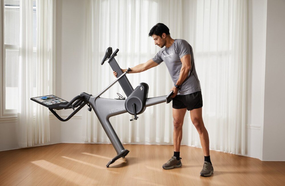 Mężczyzna w sportowym stroju ustawia rower treningowy w jasnym pomieszczeniu. Na rowerze widoczny jest ekran dotykowy oraz napis \'VELO NIX\'.
