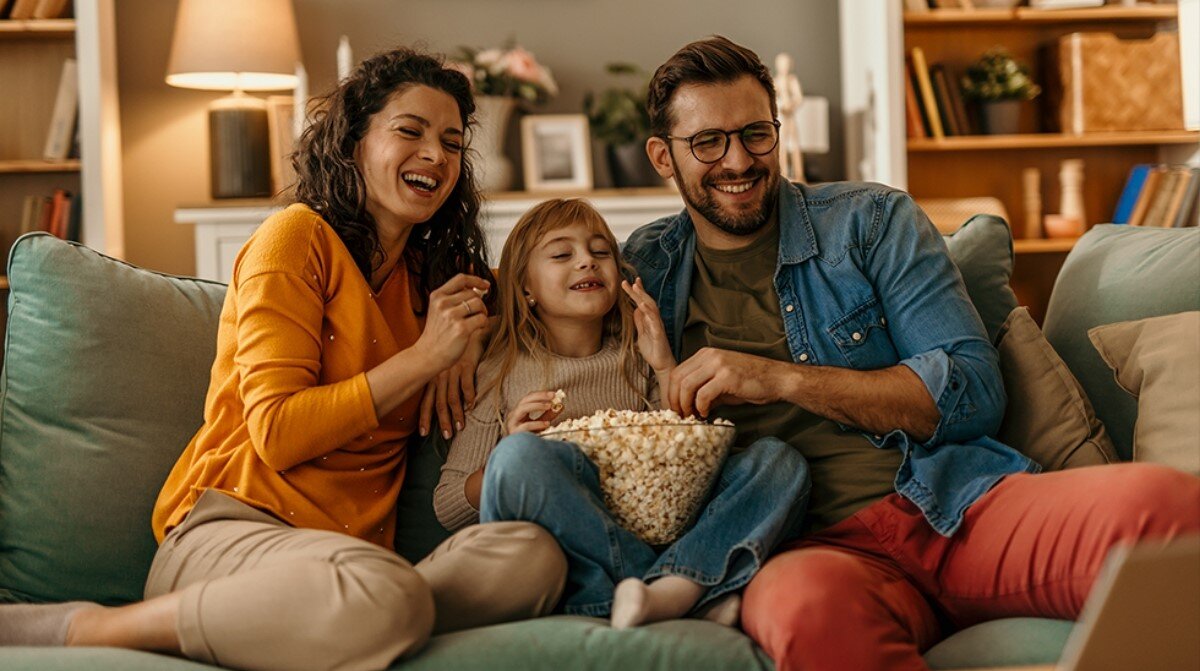 Trzy osoby siedzą na kanapie, uśmiechają się i jedzą popcorn. W tle widać lampę, półki z książkami i dekoracje.