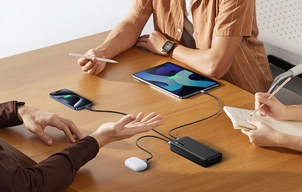 Powerbank UGREEN PB312 20000 mAh PB312 smartfony, słuchawki bezprzewodowe, smartwatche dwóch portów wyjściowych USB-A wejściowo-wyjściowego portu USB-C do trzech urządzeń - Trzy urządzenia ładowane jednocześnie z jednego powerbanku Ugreen leżącego na drewnianym stole podczas spotkania.