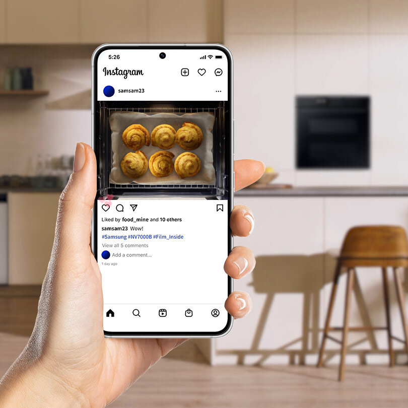 Osoba trzyma smartfon z otwartą aplikacją Instagram, na której widać zdjęcie piekarnika z pieczonymi bułeczkami. W tle znajduje się nowoczesna kuchnia z piekarnikiem w zabudowie.