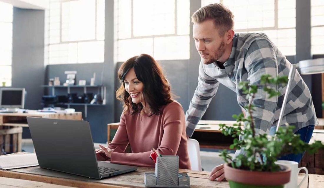 Dwie osoby siedzą przy biurku, patrząc na ekran laptopa marki Acer. Na biurku znajduje się również doniczka z rośliną i metalowy pojemnik na przybory.