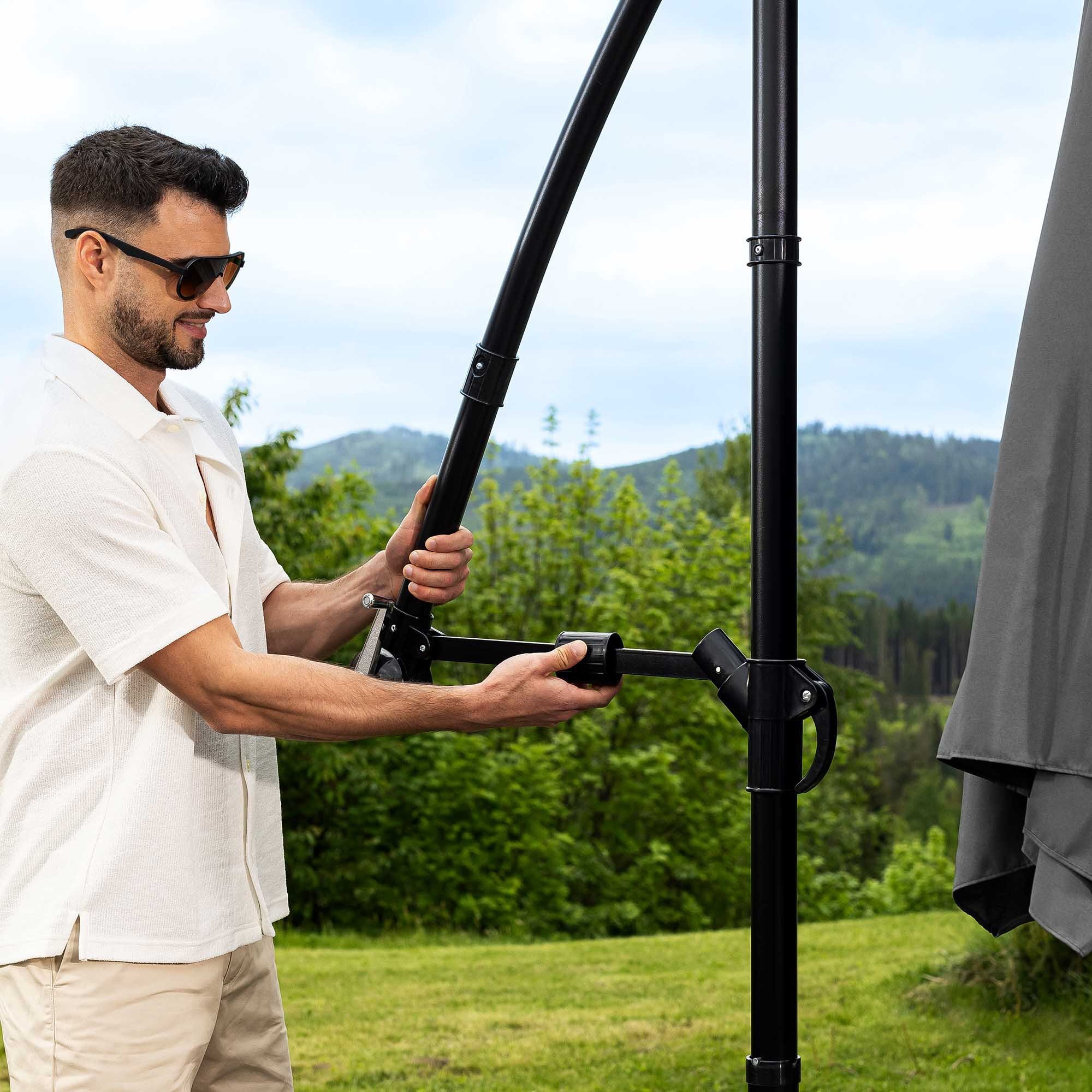 Osoba obsługuje mechanizm czarnego parasola ogrodowego na tle zielonego krajobrazu z drzewami i wzgórzami. Parasol ma szary materiał widoczny po prawej stronie.