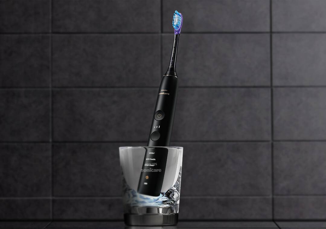 Czarna szczoteczka soniczna umieszczona w szklance na tle ciemnych płytek. Na szczoteczce widoczne są przyciski i logo Sonicare.