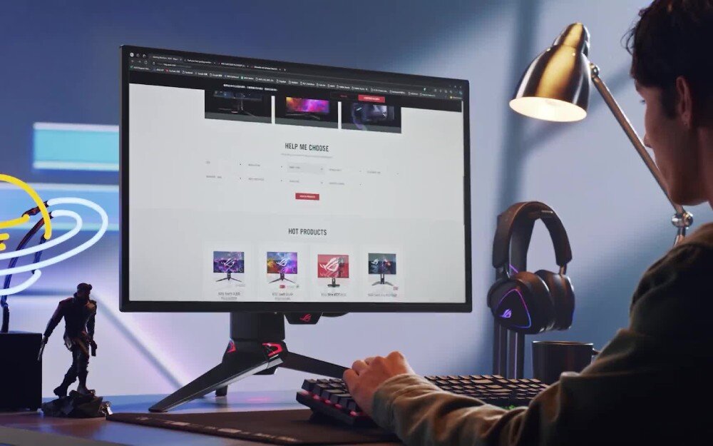 Osoba siedzi przy biurku, patrząc na monitor z wyświetloną stroną internetową. Na biurku znajdują się klawiatura, lampka i słuchawki, a w tle widać figurkę postaci.